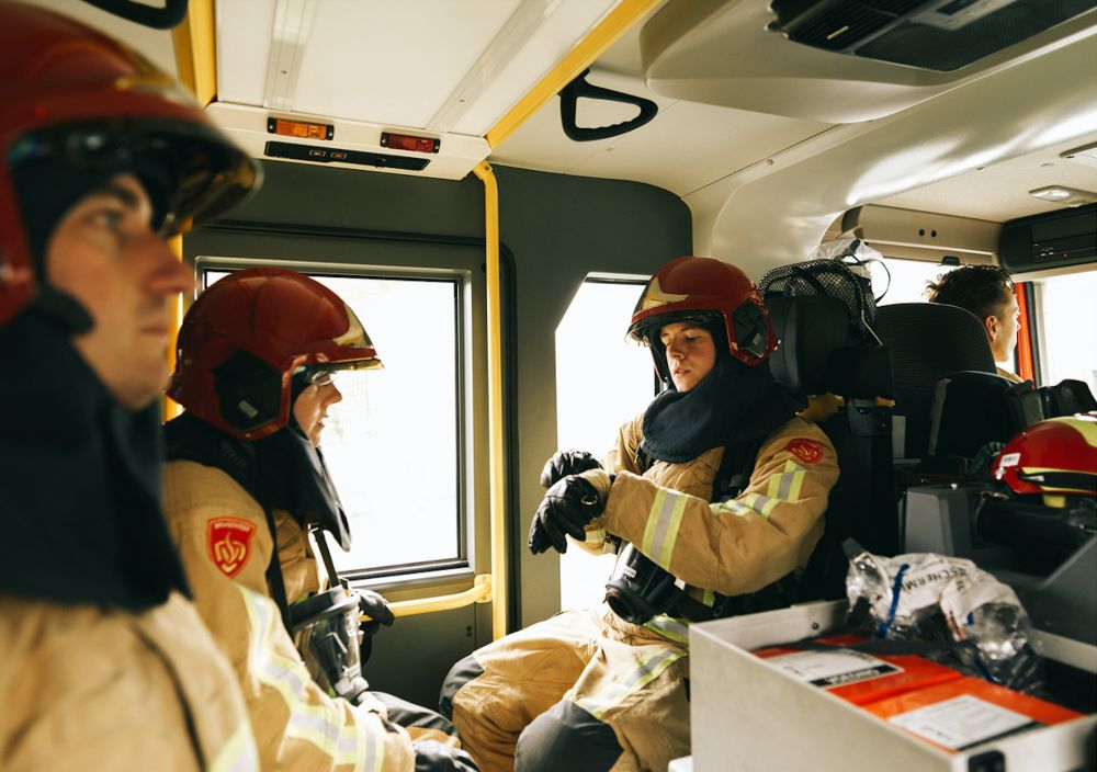 3 brandweer mannen en vrouwen achterin een brandweerwagen, een brandweervrouw trekt haar handschoen aan