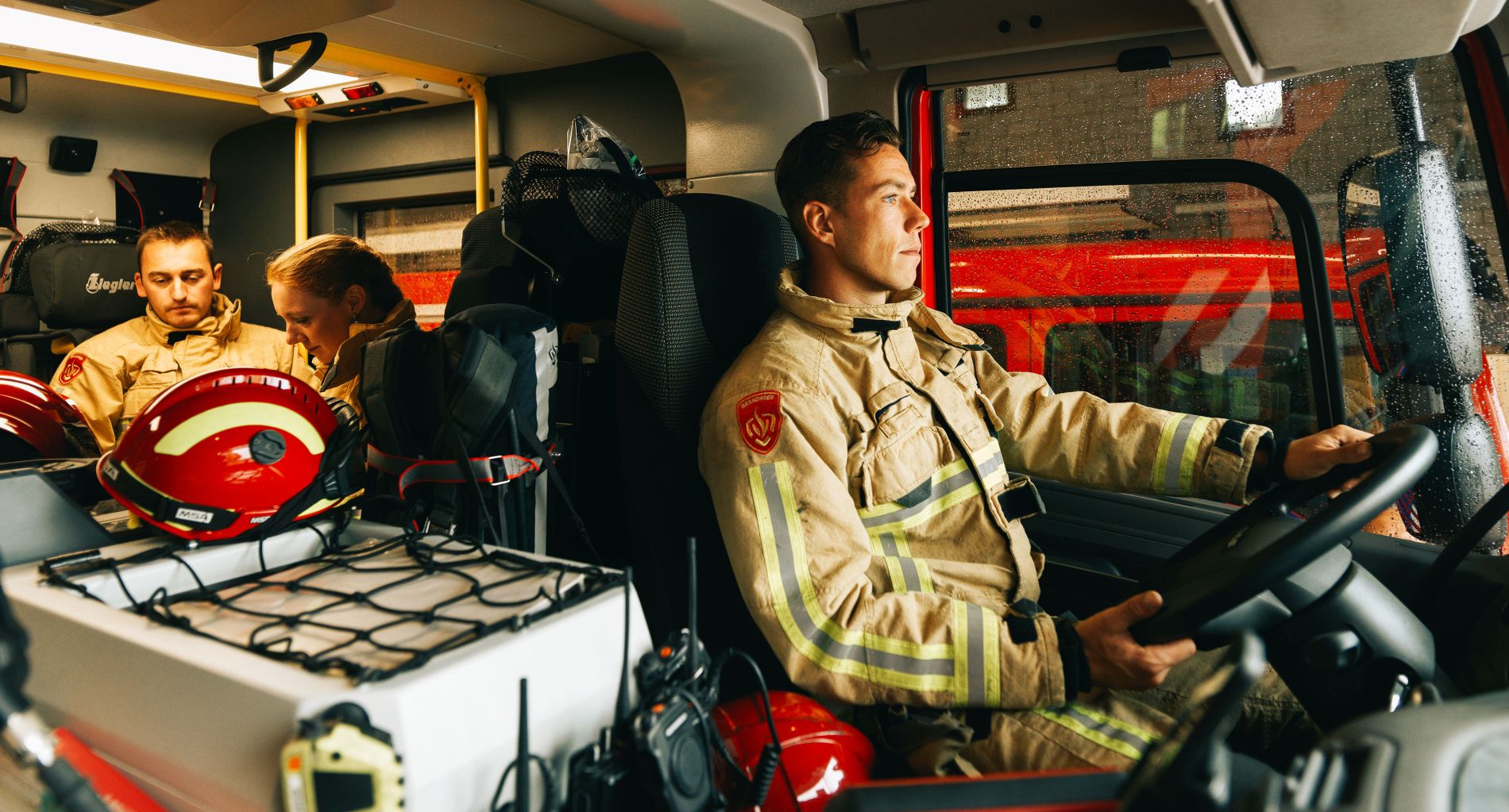 Foto in de cabine van een brandweer wagen, Brandweerman achter het stuur, achterin een brandweer-man en vrouw